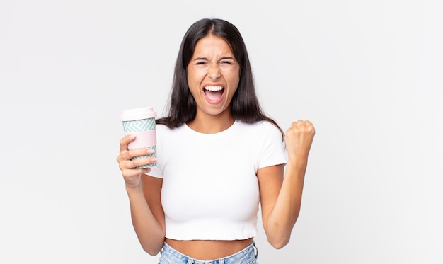 Junge hispanische Frau, die aggressiv mit einem wütenden Ausdruck schreit und einen Kaffeebehälter zum Mitnehmen hält?