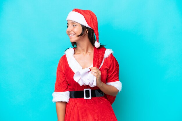 Junge hispanische Frau, die als Mama Noel verkleidet ist und einen Sack isoliert auf blauer Hintergrundseite hält