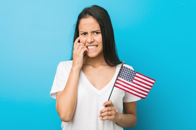 Junge hispanische Frau, die beißende Fingernägel einer Flagge Vereinigter Staaten, nervös und sehr besorgt hält.