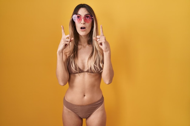 Junge hispanische Frau, die Bikini über gelbem Hintergrund trägt, erstaunt und überrascht, nach oben zu schauen und mit Fingern und erhobenen Armen zu zeigen.