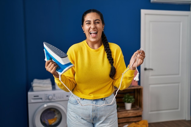 Junge hispanische Frau, die Bügeleisen in der Waschküche hält, lächelt und lacht laut, weil lustiger verrückter Witz.