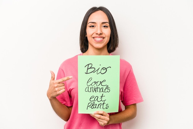 Junge hispanische Frau, die ein Bio-Plakat isoliert auf weißem Hintergrund hält, lächelt und zur Seite zeigt und etwas auf die Leerstelle zeigt