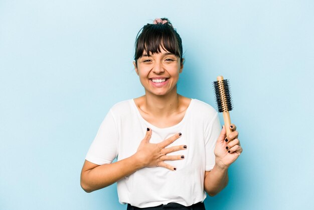 Junge hispanische Frau, die ein Bürstenhaar lokalisiert auf blauem Hintergrund hält, lacht und Spaß hat