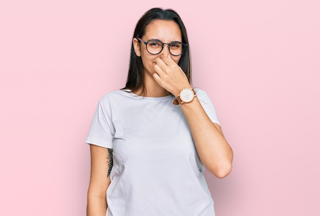 Junge hispanische Frau, die ein lässiges weißes T-Shirt trägt und etwas stinkenden und ekelhaften, unerträglichen Geruch riecht, der mit den Fingern auf der Nase den Atem anhält, schlechter Geruch