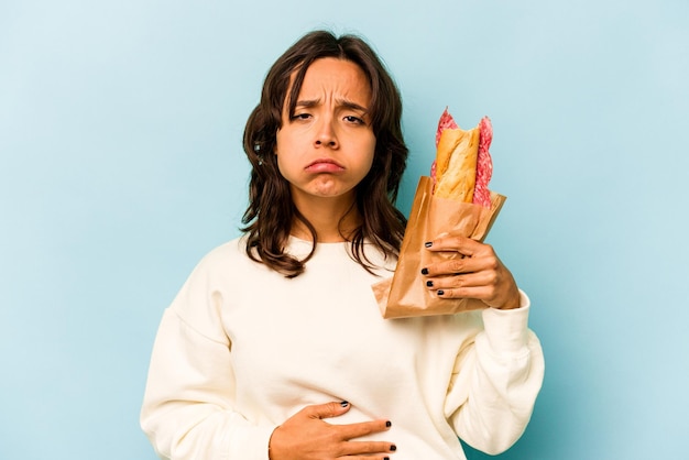 Junge hispanische Frau, die ein Sandwich isst, isoliert auf blauem Hintergrund