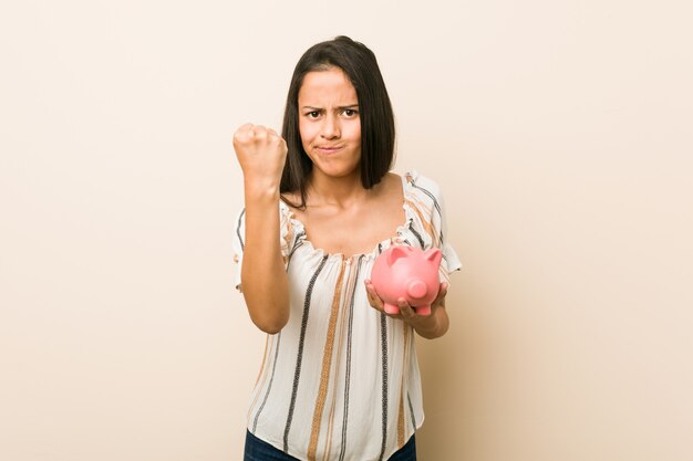 Junge hispanische Frau, die ein Sparschwein zeigt Faust zur Kamera, aggressiver Gesichtsausdruck hält.