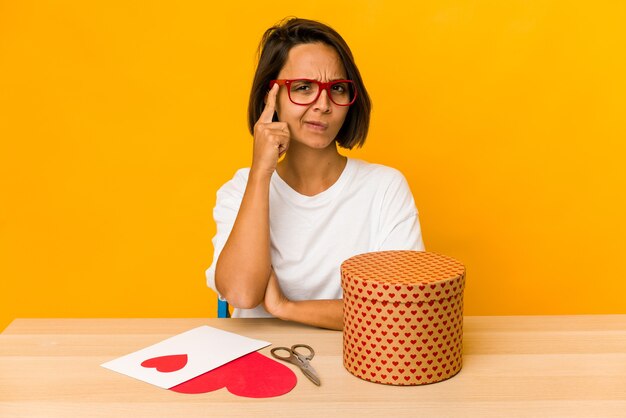 Junge hispanische Frau, die ein Valentinsgeschenk vorbereitet, lokalisiertes Zeigen des Tempels mit Finger, denkend, konzentriert auf eine Aufgabe.