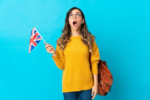 Junge hispanische Frau, die eine britische Flagge lokalisiert auf der blauen Wand hält, die oben und mit überraschtem Ausdruck schaut