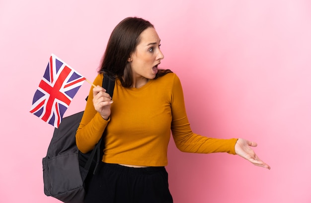 Junge hispanische Frau, die eine Flagge des Vereinigten Königreichs mit Überraschungsausdruck hält, während sie zur Seite schaut