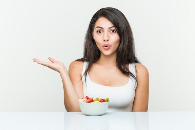 Junge hispanische Frau, die eine Obstschale isst, beeindruckte, Leerzeichen auf Handfläche haltend.