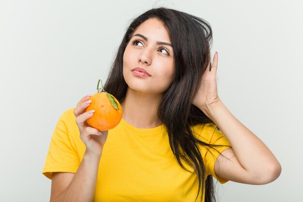 Junge hispanische Frau, die eine Orange versucht, einen Klatsch zu hören hält.