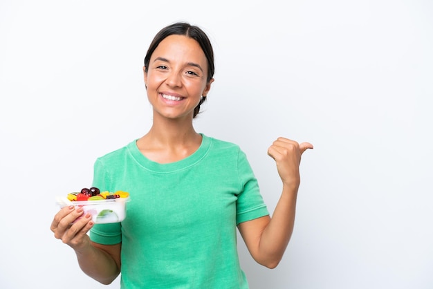 Junge hispanische Frau, die eine Schale mit Obst isoliert auf weißem Hintergrund hält, die auf die Seite zeigt, um ein Produkt zu präsentieren