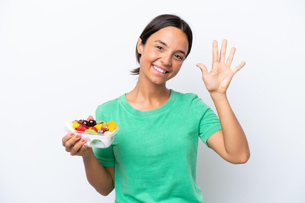 Junge hispanische Frau, die eine Schale mit Obst isoliert auf weißem Hintergrund hält und fünf mit den Fingern zählt