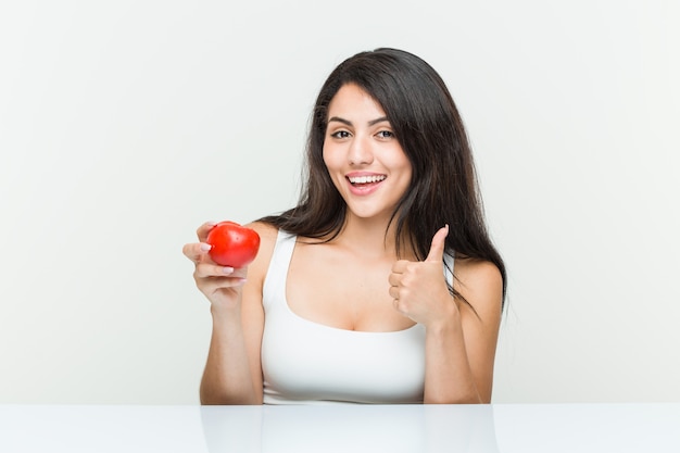 Junge hispanische Frau, die eine Tomate lächelt und Daumen hochhält