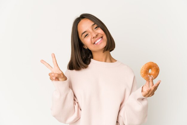 Junge hispanische Frau, die einen freudigen und sorglosen Donut isst, der ein Friedenssymbol mit Fingern zeigt.