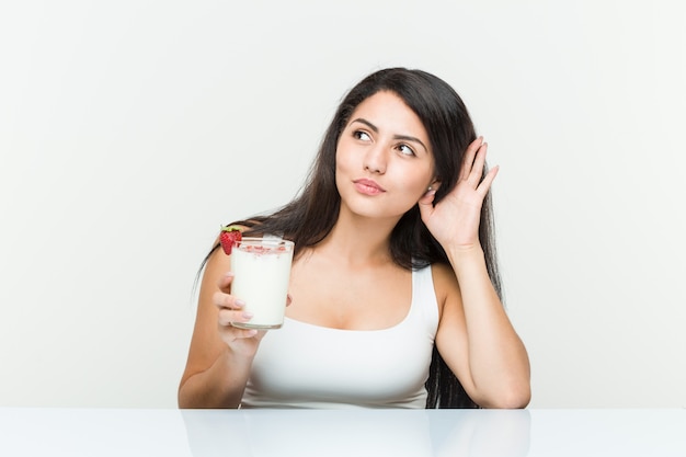 Junge hispanische Frau, die einen Smoothie hält Junge hispanische Frau, die einen Avocadotoast hält, der versucht, einen Klatsch zu hören