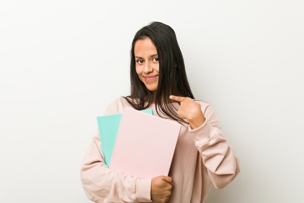 Junge hispanische Frau, die einige Notizbücher hält, die mit dem Finger auf Sie zeigen, als ob die Einladung näher kommt.