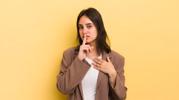 Junge hispanische Frau, die ernst und verärgert aussieht, mit dem Finger auf die Lippen gedrückt, die Schweigen fordert oder ein Geheimnis bewahrt