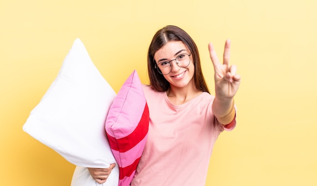 Junge hispanische Frau, die freundlich lächelt und aussieht und Nummer zwei zeigt. Pyjama- und Kissenkonzept