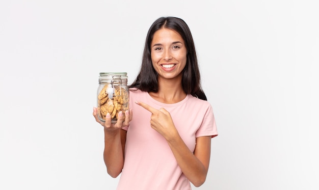 Junge hispanische Frau, die fröhlich lächelt, sich glücklich fühlt und auf die Seite zeigt und eine Glasflasche mit Keksen hält