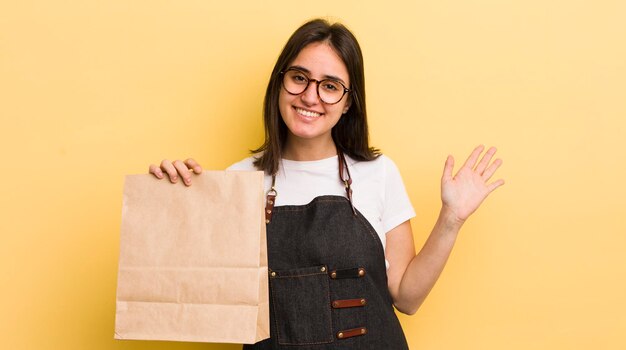 Junge hispanische Frau, die glücklich lächelt und mit der Hand winkt, begrüßt und grüßt Sie Fast-Food-Lieferkonzept