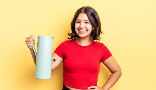 Junge hispanische Frau, die glücklich mit einer Hand auf Hüfte und überzeugt lächelt. Thermos-Konzept