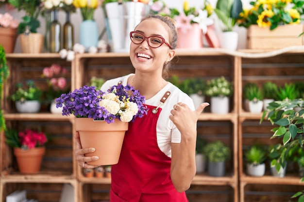 Junge hispanische Frau, die im Blumenladen arbeitet und eine Pflanze hält, die mit dem Daumen nach oben zur Seite zeigt und glücklich mit offenem Mund lächelt