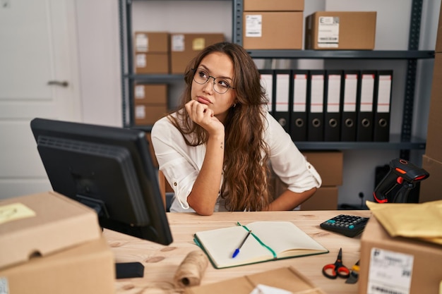 Junge hispanische Frau, die im E-Commerce-Bereich für kleine Unternehmen arbeitet, mit der Hand am Kinn, denkt über Fragen nach, nachdenklicher Gesichtsausdruck, lächelnd mit nachdenklichem Gesicht und zweifelndem Konzept