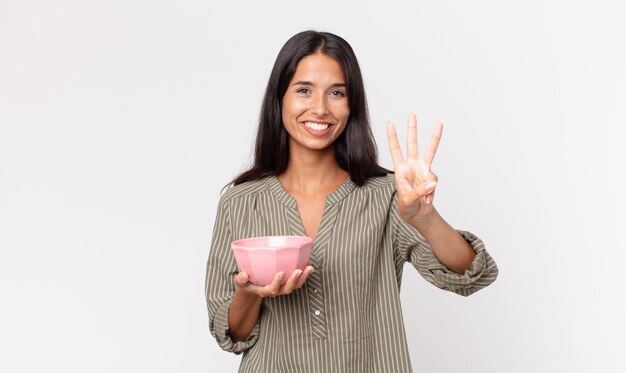 Junge hispanische Frau, die lächelt und freundlich aussieht, Nummer drei zeigt und eine leere Schüssel oder einen Topf hält