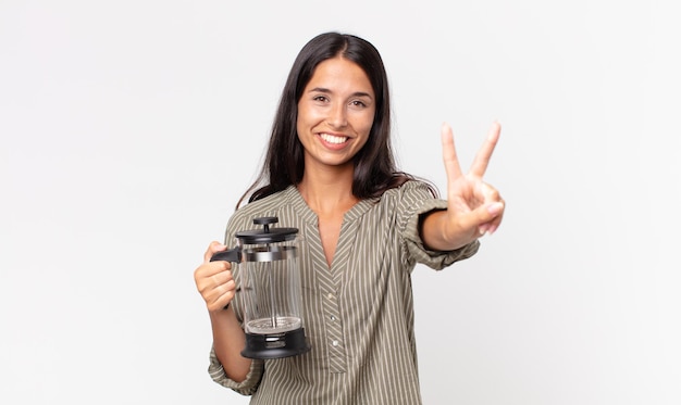 Junge hispanische Frau, die lächelt und freundlich aussieht, Nummer zwei zeigt und eine manuelle Kaffeemaschine hält