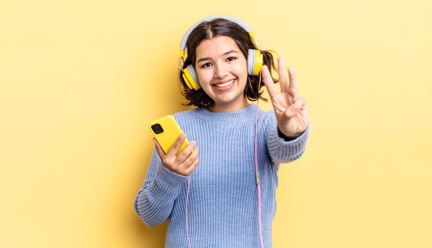 Junge hispanische Frau, die lächelt und freundlich aussieht und Kopfhörer Nummer drei und Smartphone-Konzept zeigt