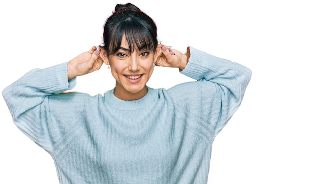 Junge hispanische Frau, die legere Kleidung trägt, lächelt und mit den Fingern an den Ohren zieht, lustiges Gesten-Vorsprechproblem