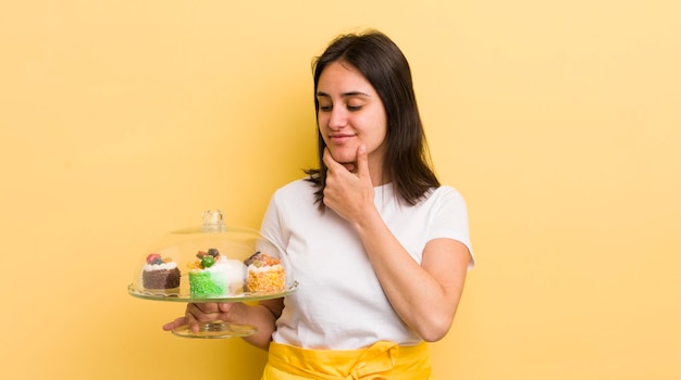 Junge hispanische Frau, die mit einem glücklichen, selbstbewussten Ausdruck lächelt, mit der Hand am Kinn, hausgemachtes Kuchenkonzept