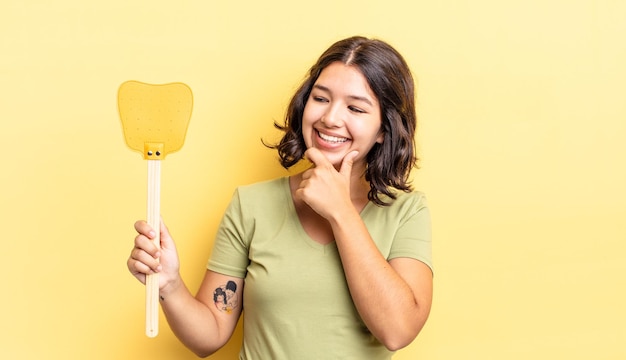 Junge hispanische Frau, die mit einem glücklichen, selbstbewussten Ausdruck mit der Hand am Kinn lächelt. Töte Fliegen Konzept