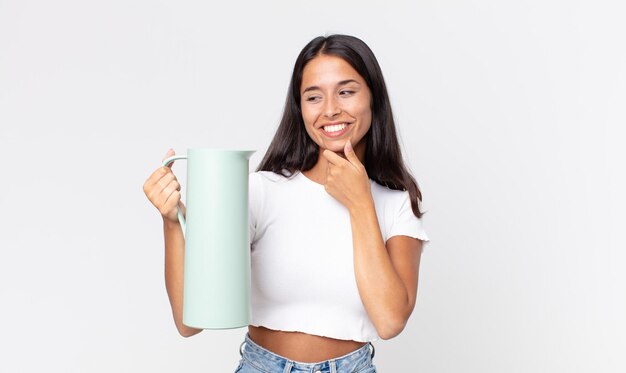 Junge hispanische Frau, die mit einem glücklichen, selbstbewussten Ausdruck mit der Hand am Kinn lächelt und eine Kaffeethermos hält