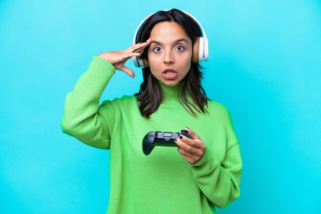 Junge hispanische Frau, die mit einem Videospiel-Controller spielt, der auf blauem Hintergrund mit überraschtem Ausdruck isoliert ist