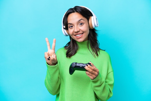 Junge hispanische Frau, die mit einem Videospiel-Controller spielt, isoliert auf blauem Hintergrund, lächelt und zeigt Victory-Zeichen