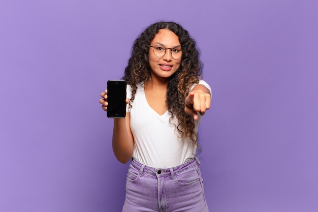 Junge hispanische Frau, die mit einem zufriedenen, selbstbewussten, freundlichen Lächeln nach vorne zeigt und Sie wählt. Smartphone-Konzept