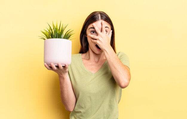 Junge hispanische Frau, die schockiert, verängstigt oder verängstigt aussieht und das Gesicht mit der Hand bedeckt. Blumentopfkonzept