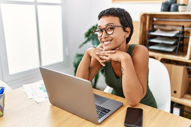 Junge hispanische Frau, die selbstbewusst lächelt und einen Laptop verwendet, der im Büro arbeitet