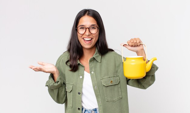 Junge hispanische Frau, die sich glücklich fühlt, überrascht, eine Lösung oder Idee zu erkennen und eine Teekanne zu halten