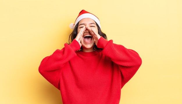 Junge hispanische Frau, die sich glücklich fühlt und mit den Händen neben dem Mund einen großen Schrei ausspricht. Weihnachtskonzept