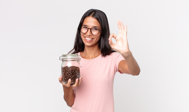 Junge hispanische Frau, die sich glücklich fühlt, Zustimmung mit okayer Geste zeigt und eine Kaffeebohnenflasche hält