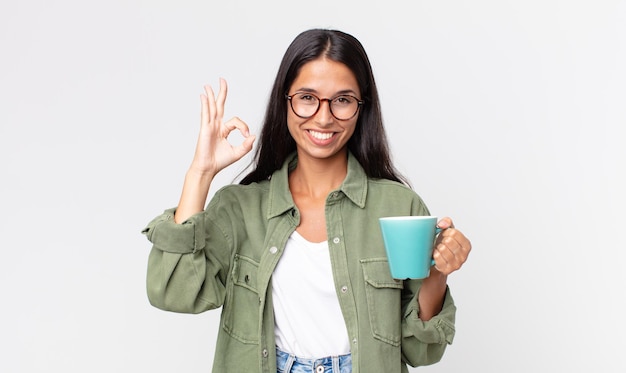 Junge hispanische Frau, die sich glücklich fühlt, Zustimmung mit okayer Geste zeigt und eine Kaffeetasse hält