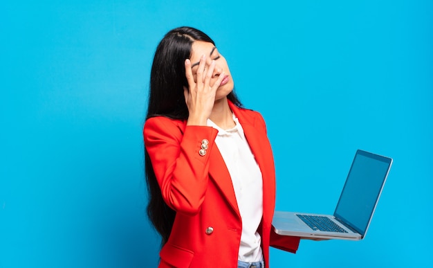 Junge hispanische Frau, die sich nach einer ermüdenden, langweiligen und mühsamen Aufgabe gelangweilt, frustriert und schläfrig fühlt und das Gesicht mit der Hand hält. Laptop-Konzept