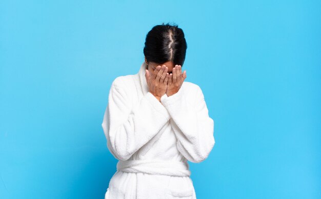Junge hispanische Frau, die sich traurig, frustriert, nervös und depressiv fühlt, Gesicht mit beiden Händen bedeckt und weint. Bademantel-Konzept