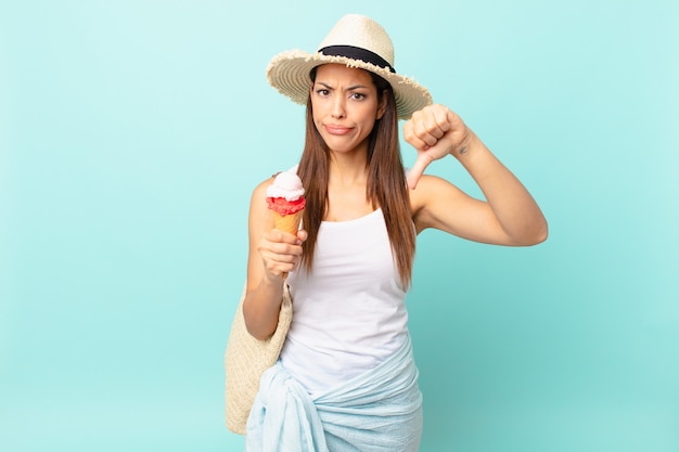 Junge hispanische Frau, die sich überquert, Daumen nach unten zeigt und ein Eis hält. Sommerkonzept