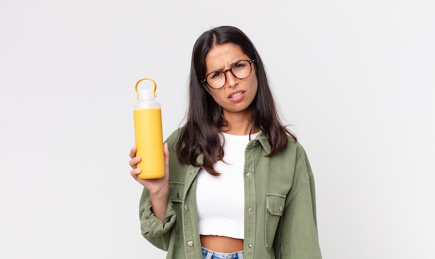 Junge hispanische Frau, die sich verwirrt und verwirrt fühlt und eine Kaffee-Thermoskanne hält