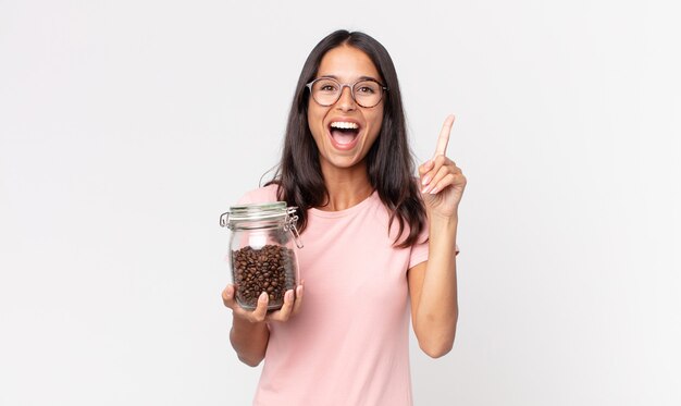 Junge hispanische Frau, die sich wie ein glückliches und aufgeregtes Genie fühlt, nachdem sie eine Idee realisiert und eine Kaffeebohnenflasche in der Hand hält