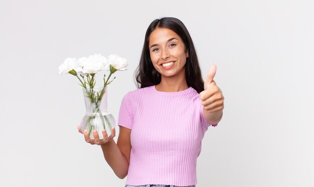 Junge hispanische Frau, die stolz ist und positiv mit Daumen nach oben lächelt und dekorative Blumen hält